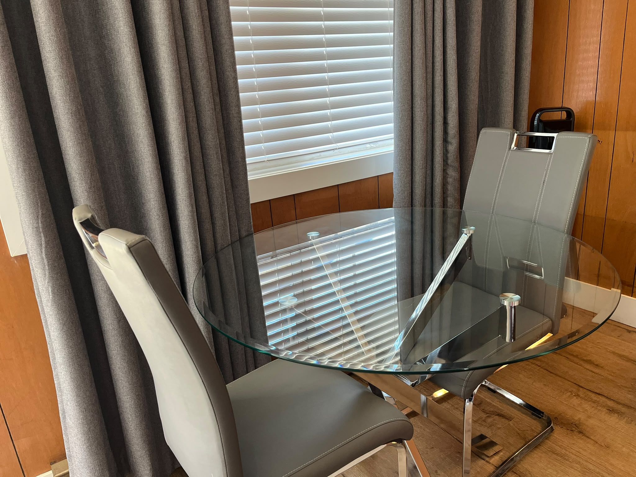 A modern room with a glass table and chairs, a window with blinds, and a wooden floor.