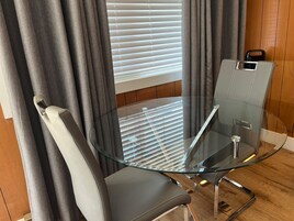Quarto moderno com mesa e cadeiras de vidro, janela com persianas e piso de madeira.