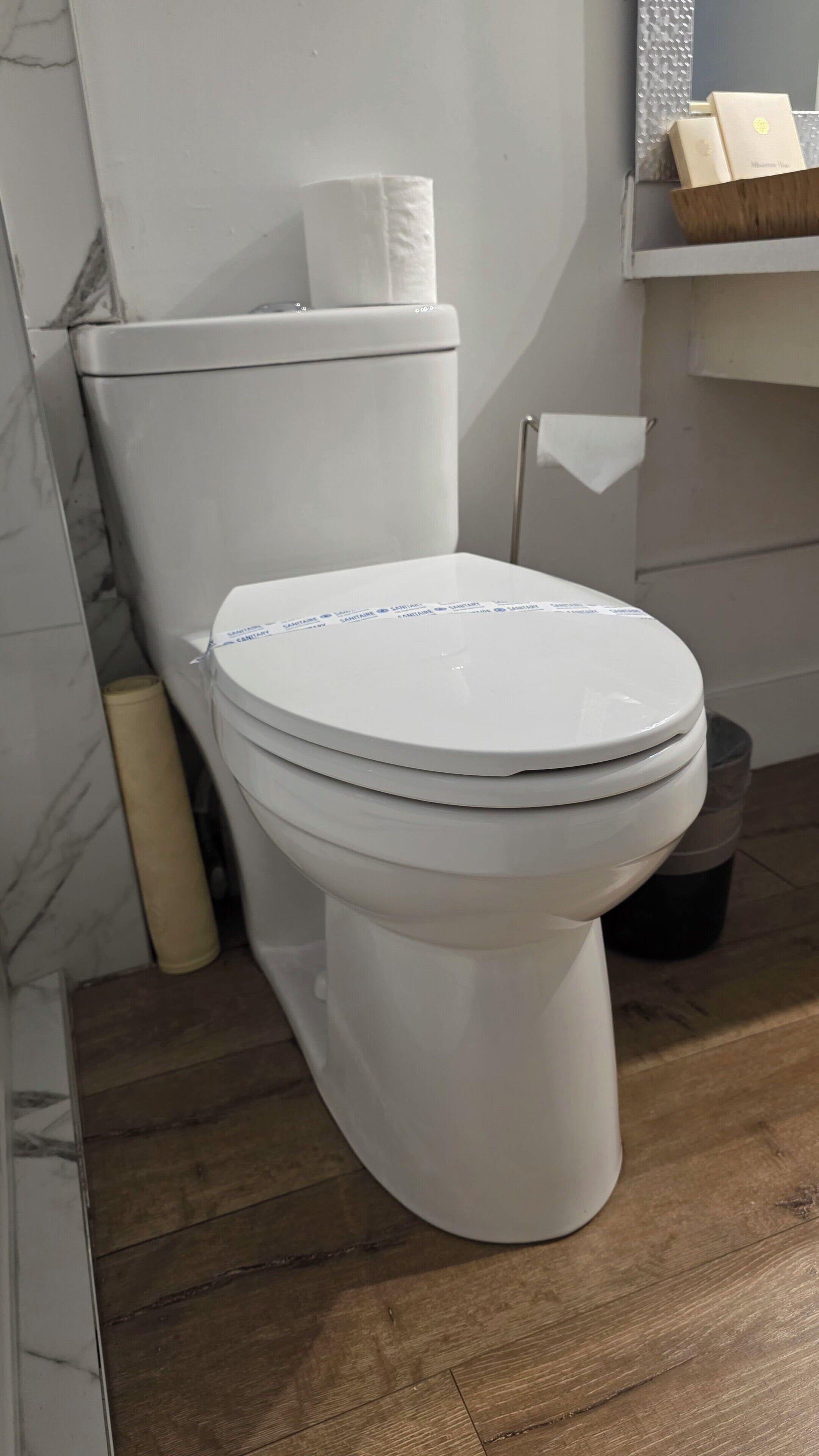 Inodoro blanco con etiqueta azul, pared de mármol y suelo de madera.