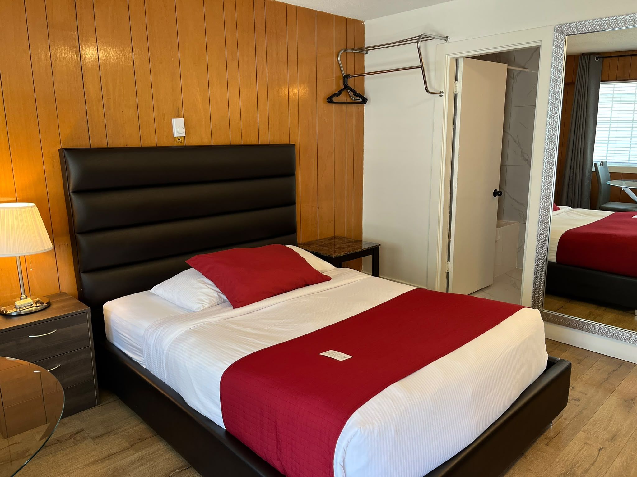 A hotel room with a bed, a nightstand, a lamp, a mirror, and a door leading to another room.
