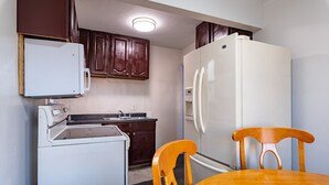 Een kleine keuken met een fornuis, spoelbak en koelkast, houten kastjes en een eettafel met stoelen.