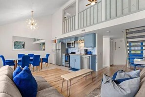 Amplia sala de estar con techos altos, una mesa de comedor, sillas azules, un sofá con cojines azules y una zona de cocina con armarios azules.