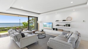 Un salon moderne doté d’une grande fenêtre donnant sur une plage, d’un ensemble de canapés, d’une table basse et d’un téléviseur fixé au mur.
