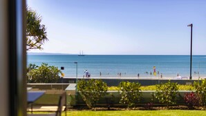 Une vue sur la plage, avec un ciel dégagé, un voilier à l’horizon et une promenade bordée de personnes et de parasols.