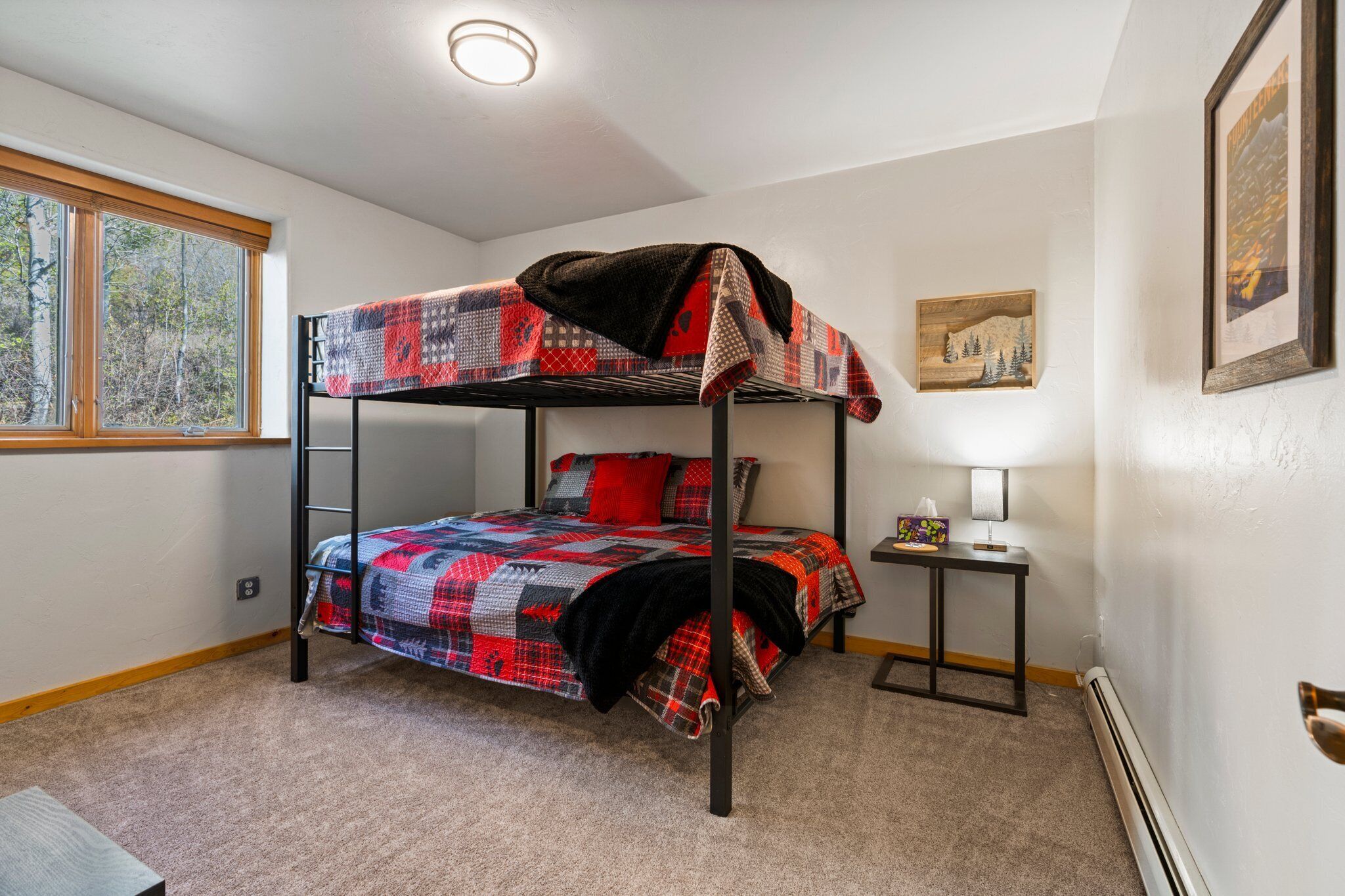 A bunk bed with a red and black checkered bedspread, a small bedside table with a lamp, and a window with a view of trees.