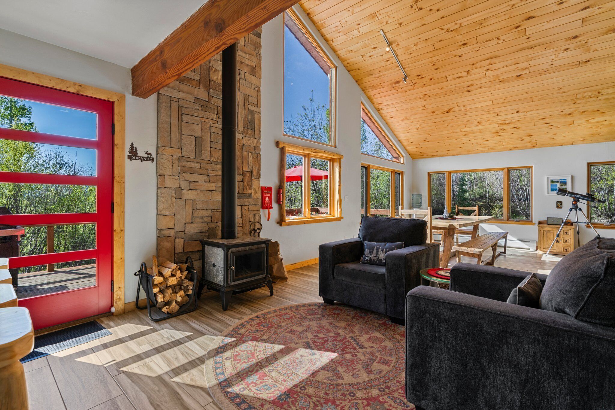 A cozy living room with a stone fireplace, large windows, and a high wooden ceiling.