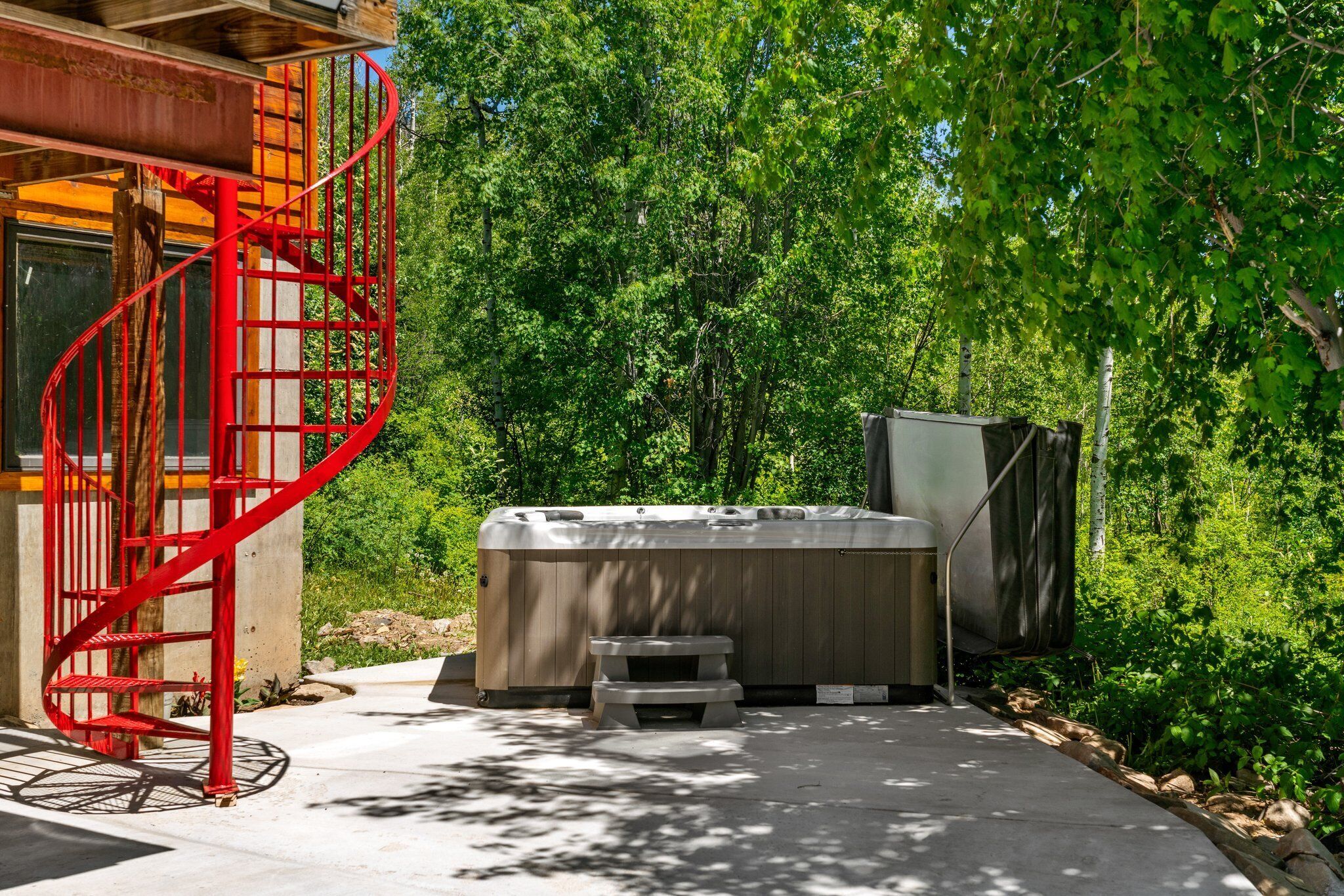 Un bain à remous avec un revêtement extérieur en bois, un escalier en colimaçon rouge et une terrasse en béton entourée de verdure.