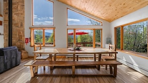 A spacious dining area with a wooden table and benches, large windows offering views of the outdoors, and a stone wall on one side. - Secluded Getaway Near Purgatory - Luxurious Hot Tub, Idylic 360° Views, Pets OK! (Durango)