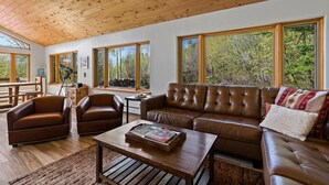 A spacious living room with a wooden ceiling, leather sofas, a coffee table, and large windows offering a view of trees. - Secluded Getaway Near Purgatory - Luxurious Hot Tub, Idylic 360° Views, Pets OK! (Durango)