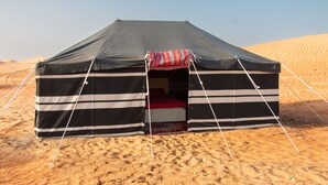 A black and white striped tent with a red and white patterned entrance mat, set in a desert with sand dunes in the background.