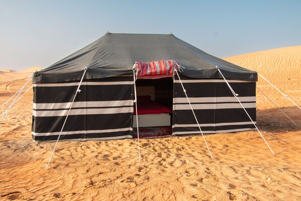 A black and white striped tent with a red and white patterned entrance mat, set in a desert with sand dunes in the background.