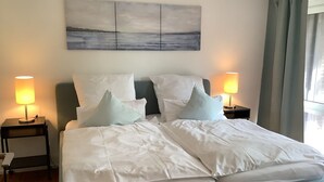 Una cama bien hecha con sábanas blancas y dos almohadas, flanqueada por mesitas de noche con lámparas, en una habitación con un cuadro abstracto grande en la pared y vistas al exterior a través de una ventana.