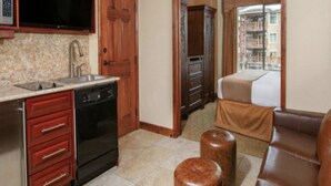 A compact hotel room with a kitchenette, a bed, a window with a view of buildings, and a ceiling fan.