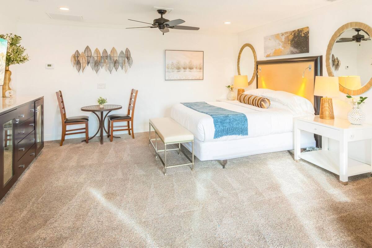 A hotel room with a bed, bedside table, lamp, mirror, artwork, and a desk with chairs.