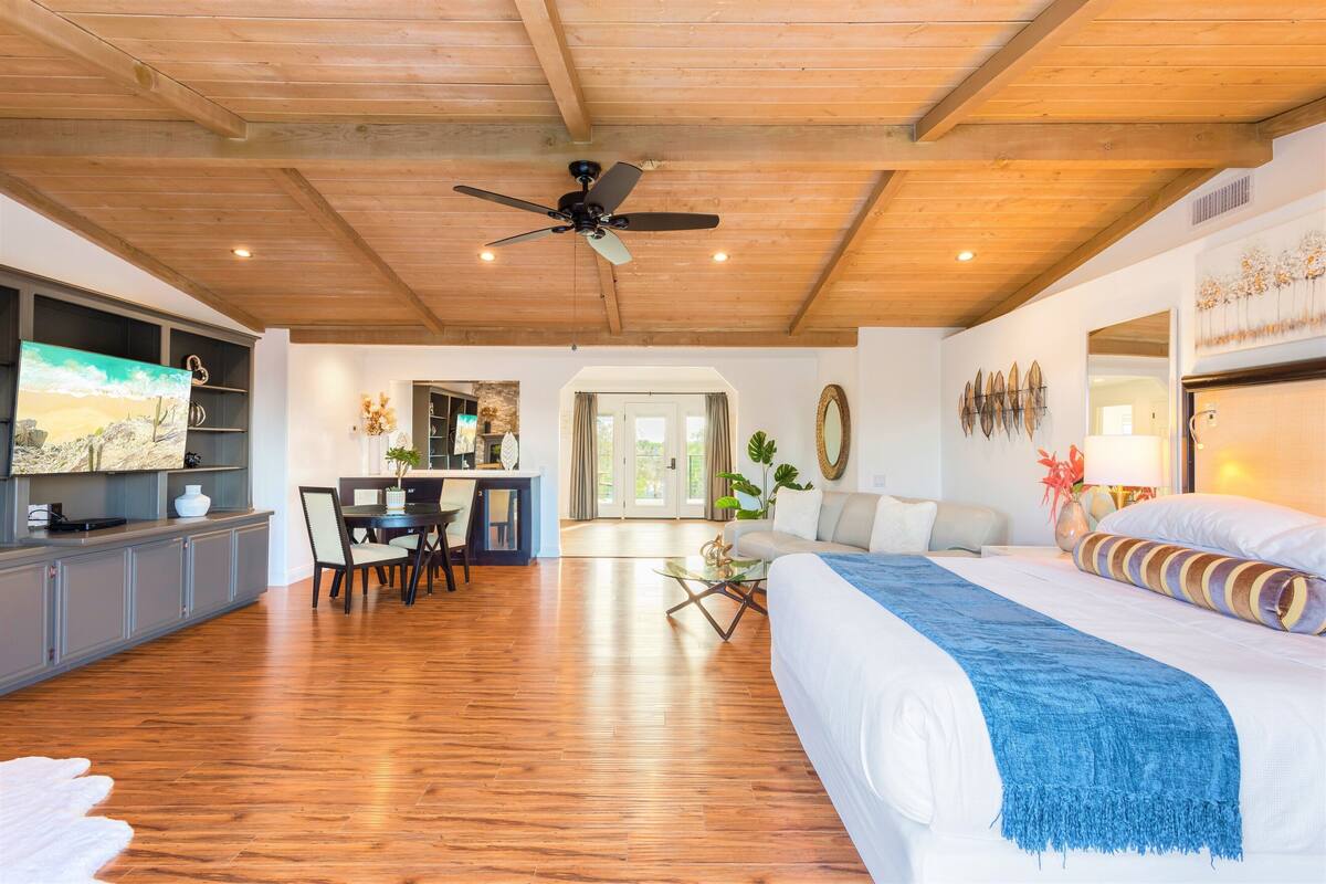 A spacious room with a wooden ceiling, a bed with a blue throw, a dining area, and a TV on the left wall.