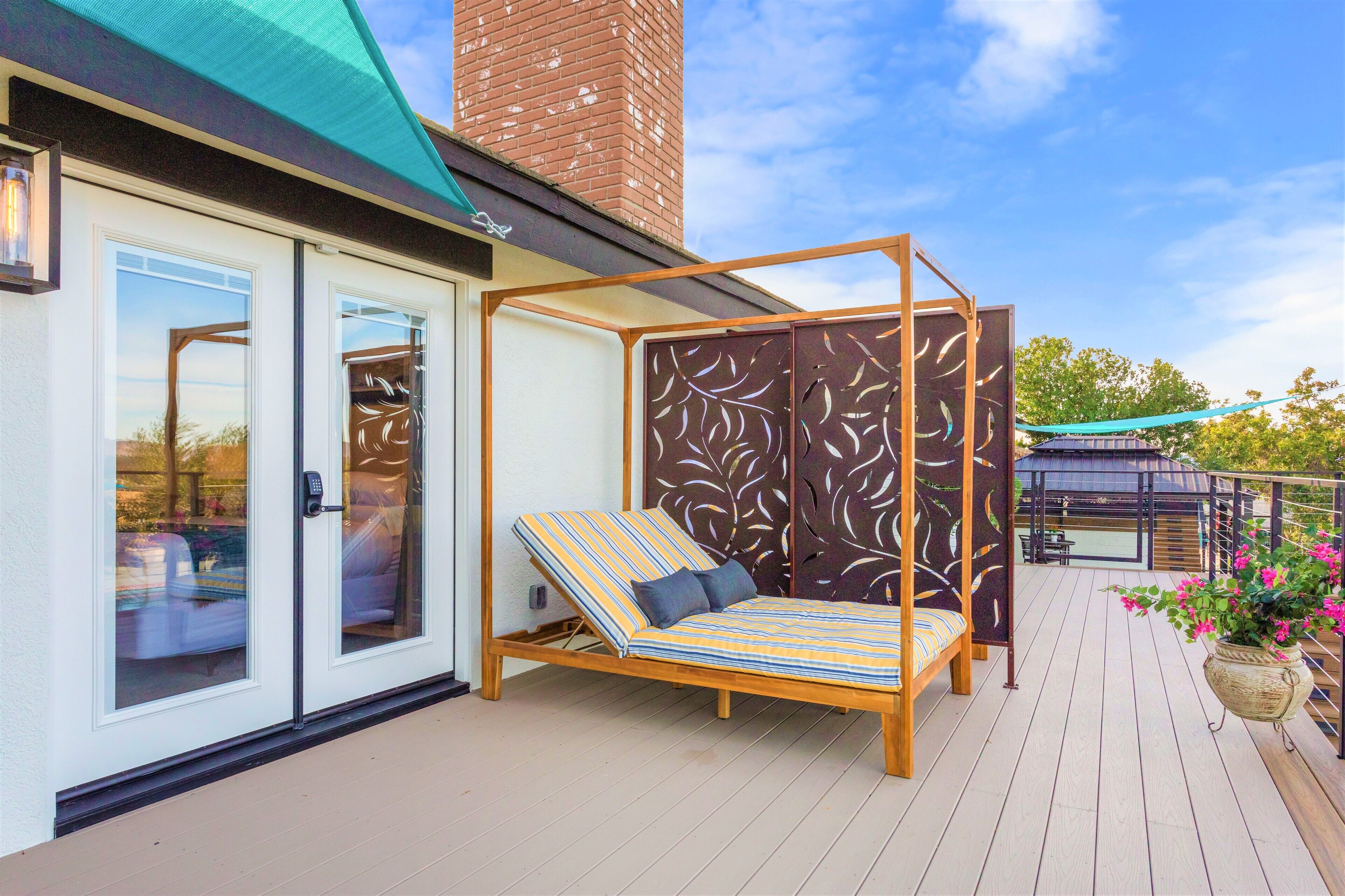 Une chaise longue avec des coussins à rayures, placée sur une terrasse en bois, avec une porte-fenêtre coulissante et un panneau décoratif orné de motifs de feuilles.