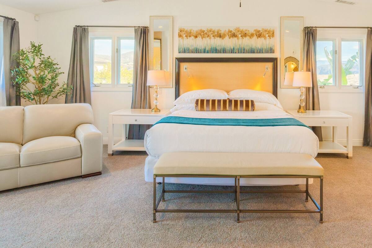 A hotel room with a bed, a bench, a sofa, two bedside tables with lamps, a large mirror, and a potted plant.