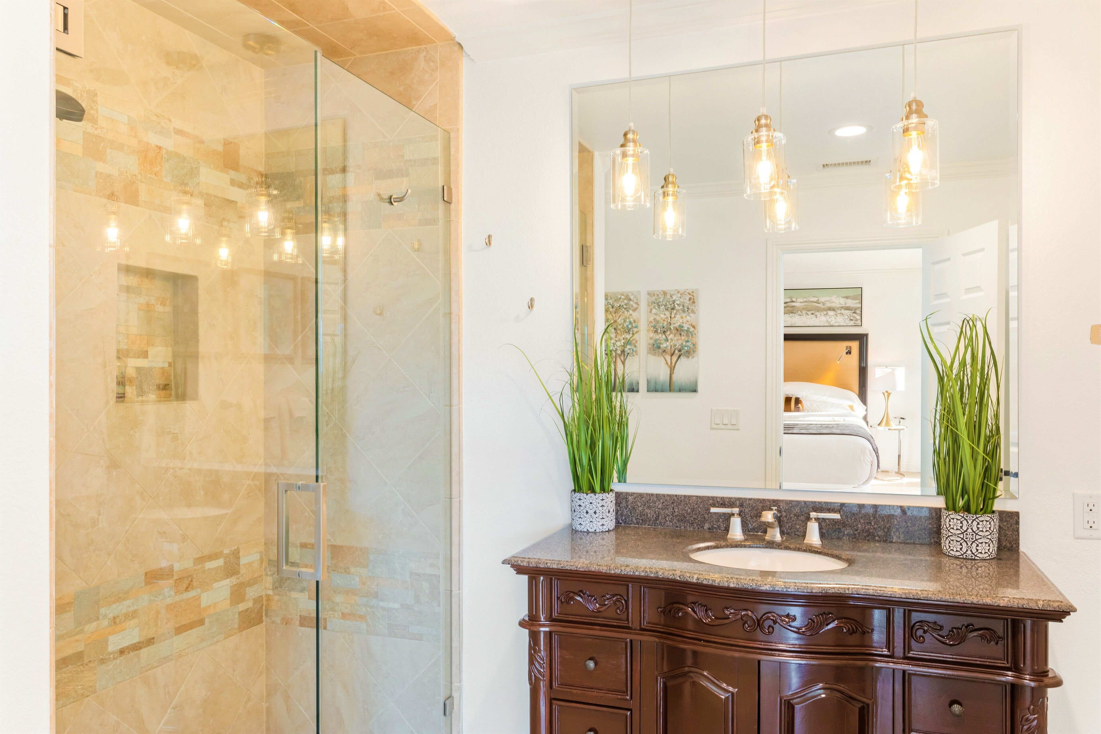 A bathroom with a glass shower enclosure, a sink with a marble countertop, and a large mirror with pendant lights.