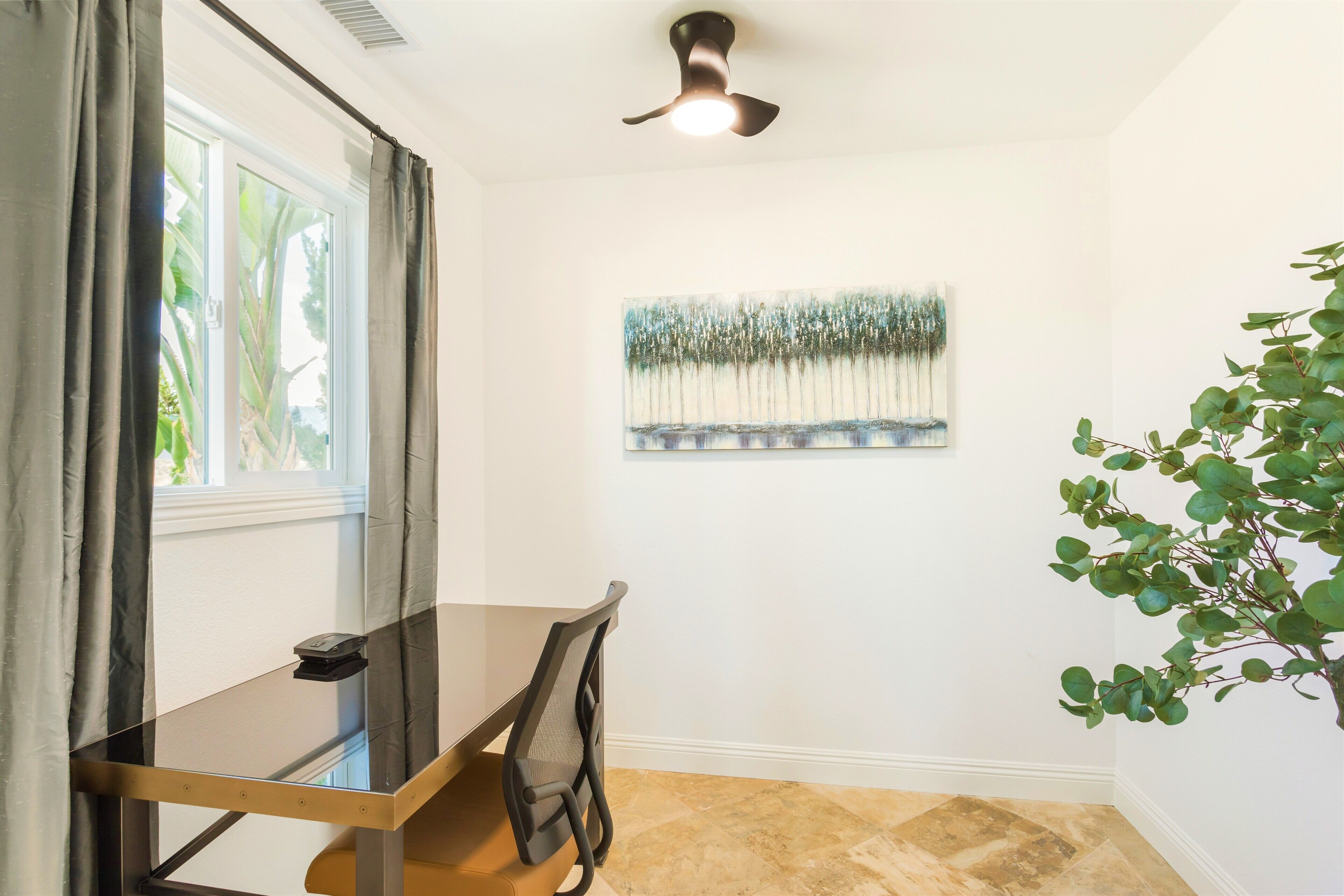 Un coin repas comprenant une table en verre, une chaise, un ventilateur de plafond, une fenêtre avec des rideaux et un tableau accroché au mur.