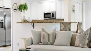 Un salon moderne comprenant un canapé, deux petites tables rondes, une chaise en bois et un coin cuisine avec des meubles blancs et un four à micro-ondes.