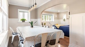 Un coin repas avec une table rectangulaire, des chaises blanches et une nappe à motifs. Des plantes en pot sont posées sur le rebord de la fenêtre, et un canapé bleu se trouve à l’arrière-plan.