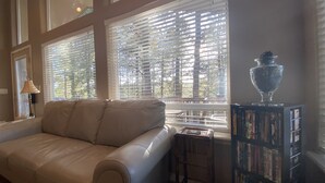 A living room with a beige sofa, a small table with a lamp, and a bookshelf filled with books. Large windows with blinds allow natural light to enter. - Home in the Pines • Bathroom for Every Bedroom (Flagstaff)