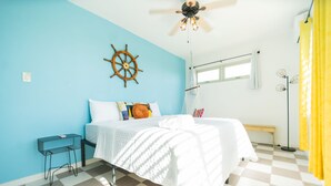 Um quarto com chão quadriculado, uma cama com roupa de cama branca, um leme de madeira na parede, uma pequena mesa e um candeeiro suspenso.