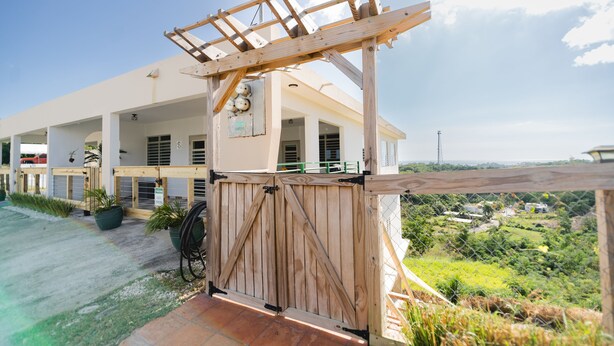 Um portão de madeira com pérgola, dando acesso a um edifício com varandas e vista para uma paisagem verde.