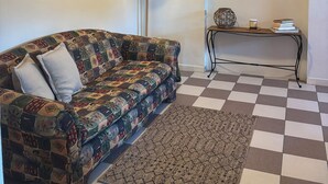 Uma sala de estar com piso xadrez, um sofá estampado, um ventilador de teto, um abajur fixado na parede, um quadro emoldurado e um espelho decorativo.