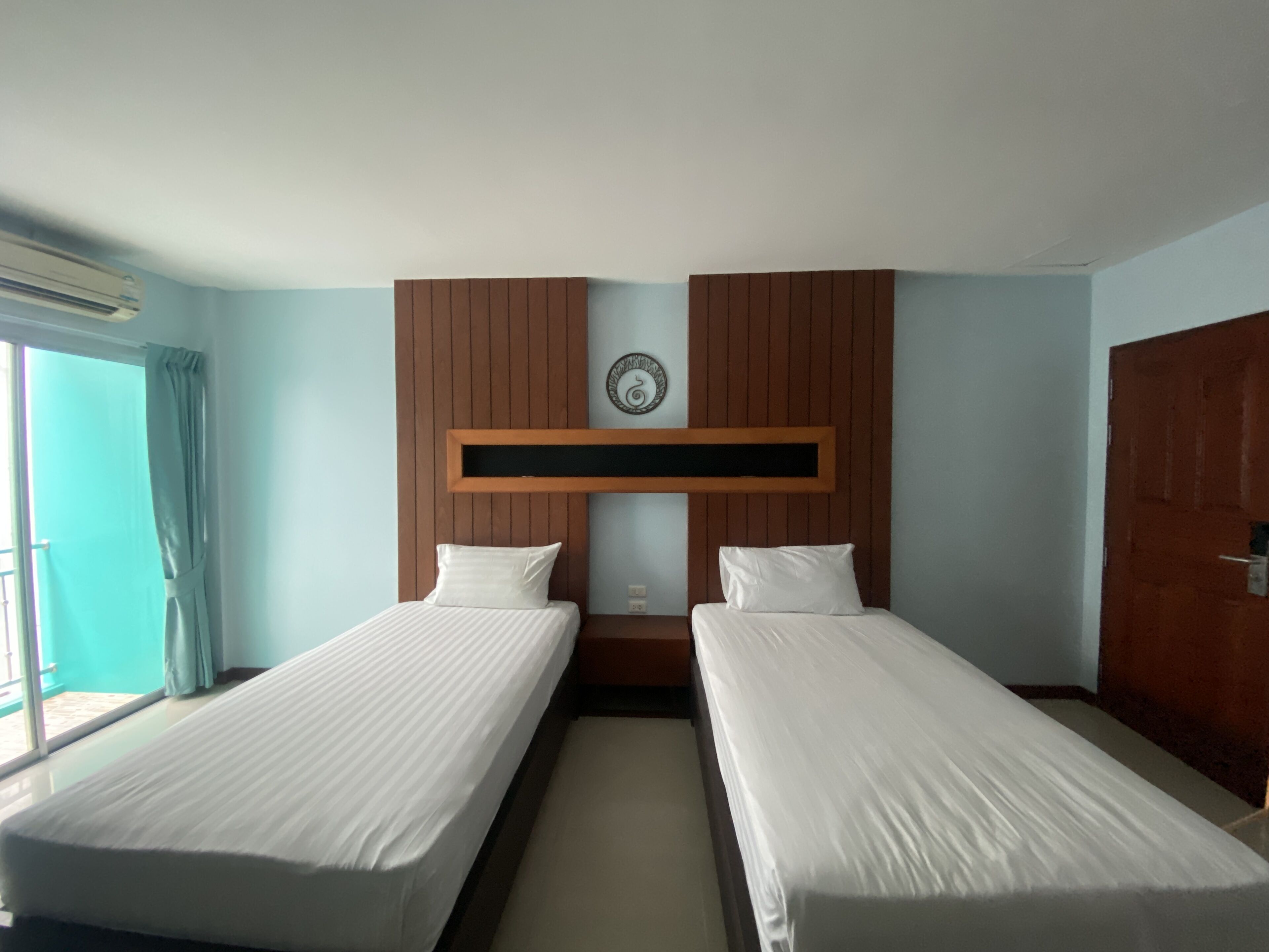 A hotel room with two single beds, a wooden headboard, a small wall clock, and a window with curtains.
