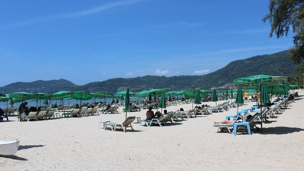 A beach with rows of sun loungers and umbrellas, a boat on the sand, and mountains in the background. - Beachwalk Patong (Patong)