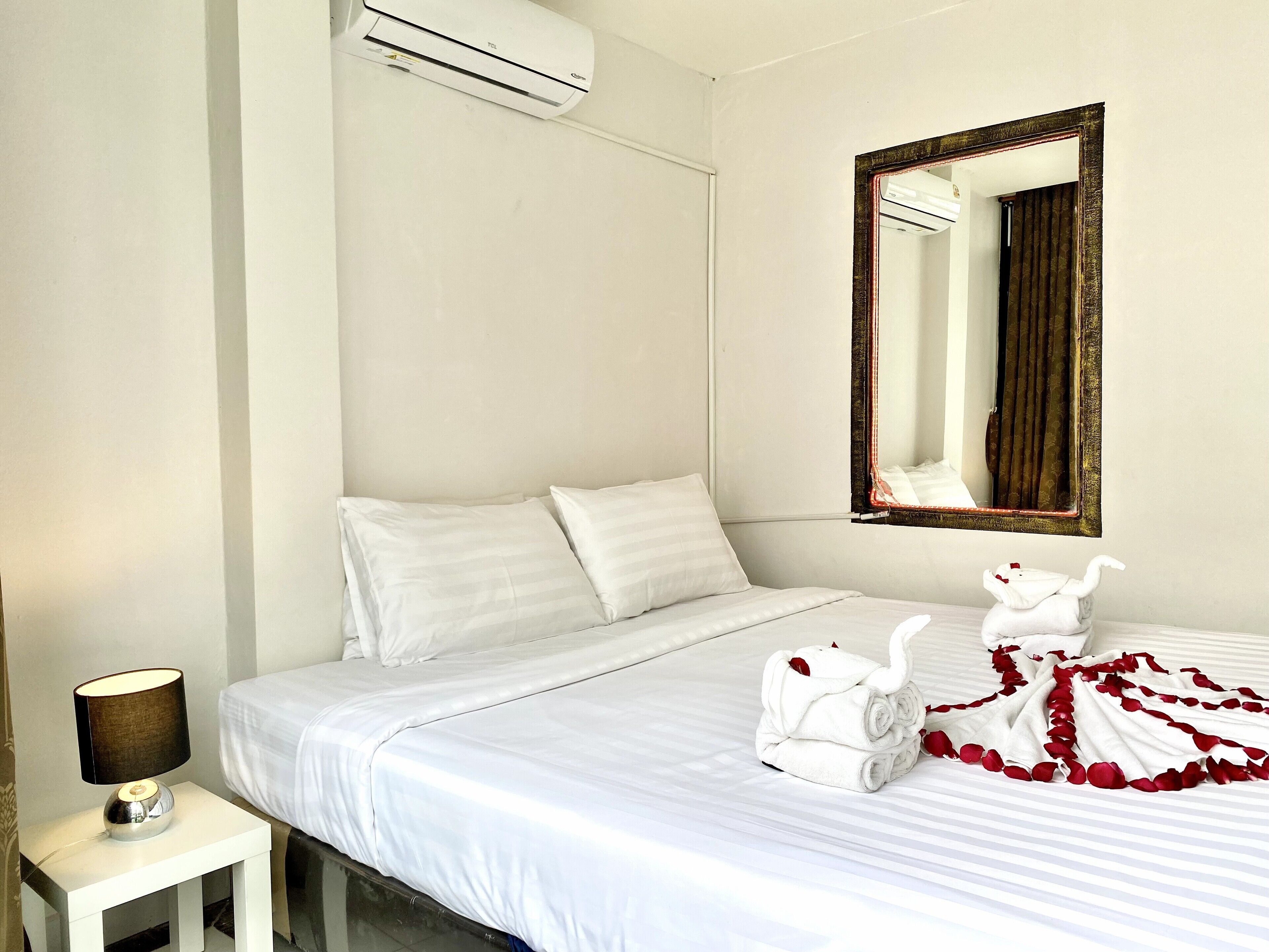 A hotel room with a bed, a nightstand with a lamp, an air conditioning unit, and a mirror with a decorative frame.