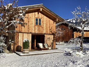A wooden cabin with a large window, a porch with a chair, and a snow-covered landscape. - Chalet am Birnbaum, entire house, 140 m² for 2–6 people, 2 bedrooms, 2 bathrooms, sauna (Unterwössen)