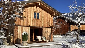 A wooden cabin with a large open door, a small porch with a chair, and a snow-covered landscape. - Chalet am Quittenbaum, whole house, 140 sqm for 2-6 persons, 2 bedrooms, 2 bathrooms, sauna (Unterwössen)