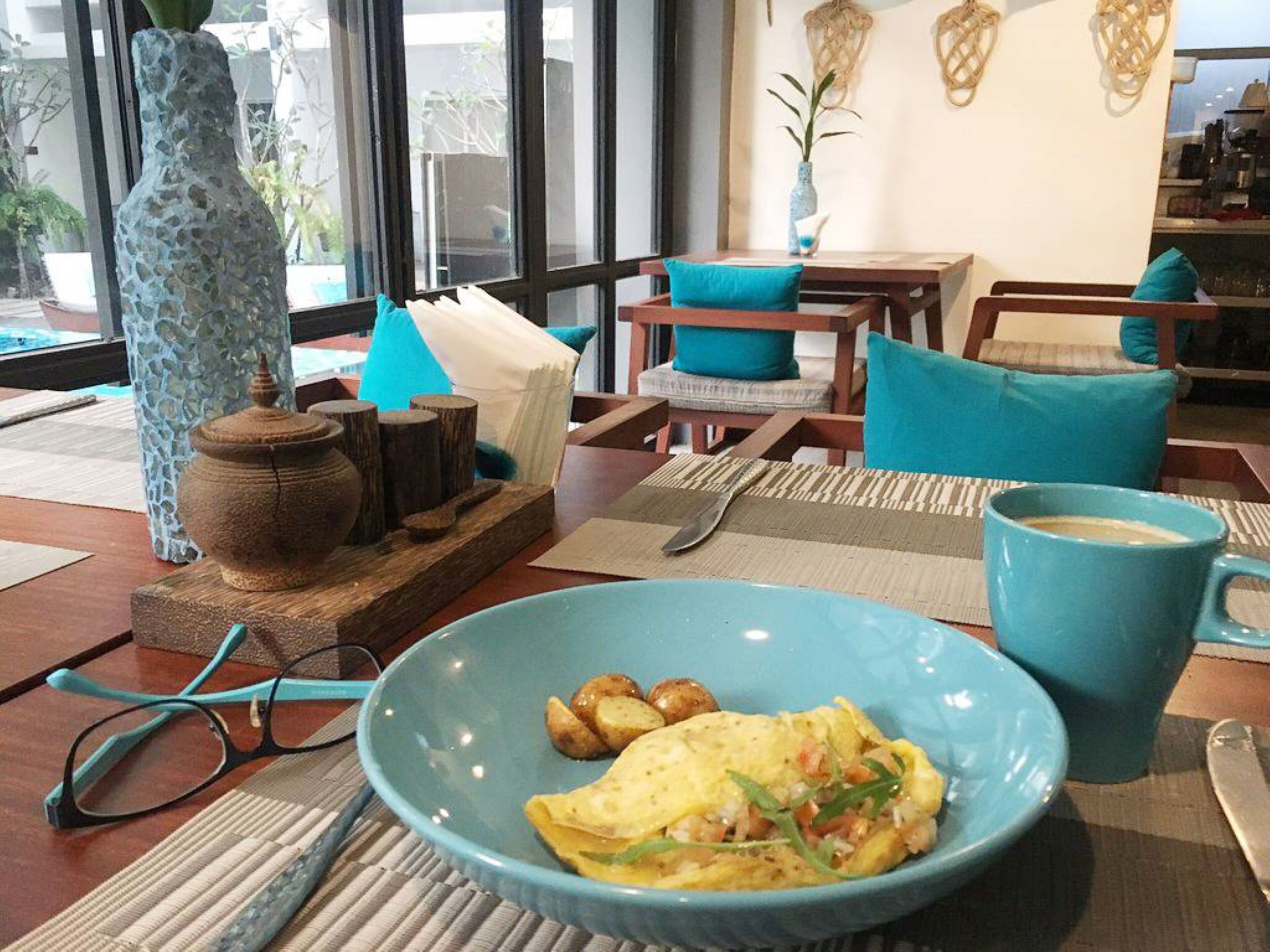 Comedor con una mesa dispuesta para dos personas, mostrando un plato azul con tortilla de patatas, una taza azul y un par de vasos sobre un tapete tejido.