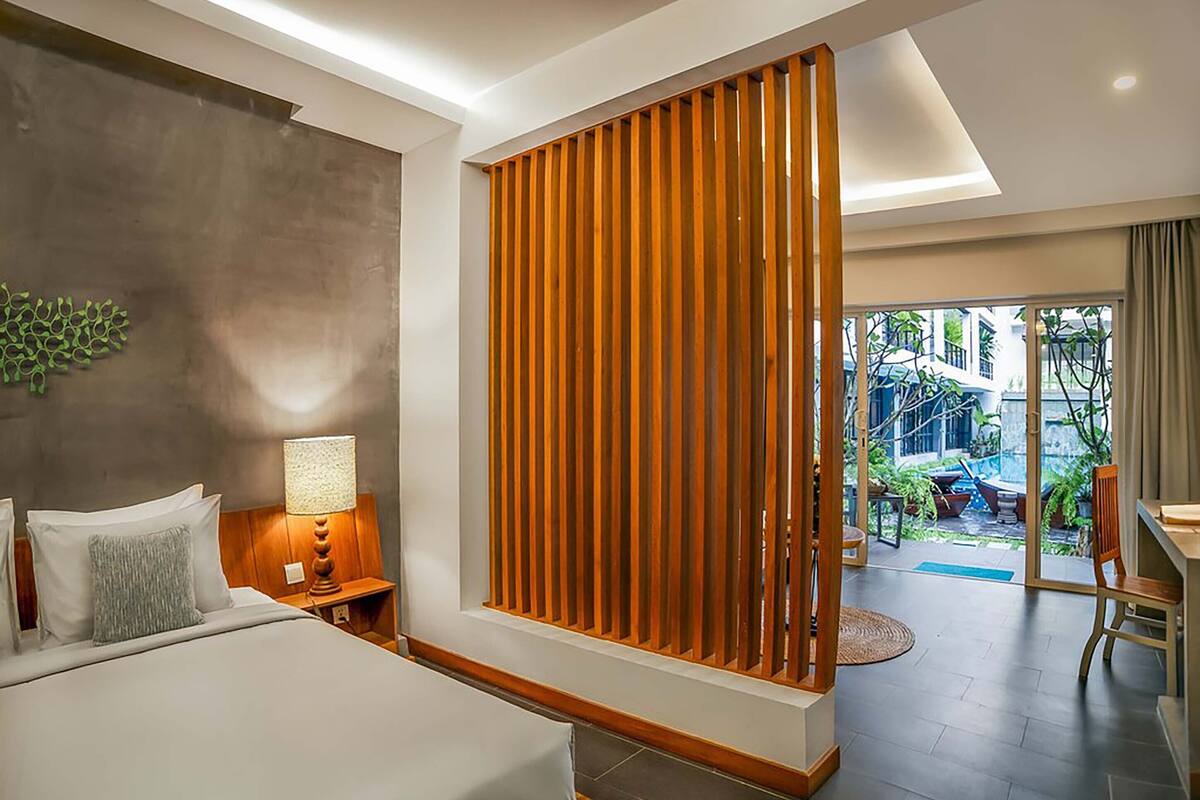 A modern hotel room with a large bed, a wooden partition, a desk, and a view of the outdoor garden.
