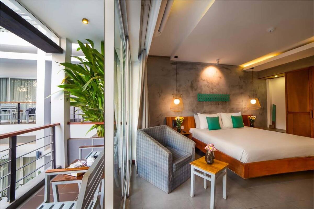 A modern bedroom with a large bed, a small seating area, a balcony with a table, and a large plant.
