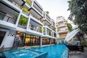 Et moderne hotel med en central swimmingpool, flere altaner med planter og en restaurant med udendørs siddepladser.