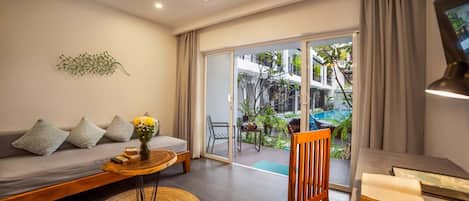 A modern living room with a sofa, a wooden coffee table, and a chair. There is a view of a balcony with plants and a pool.