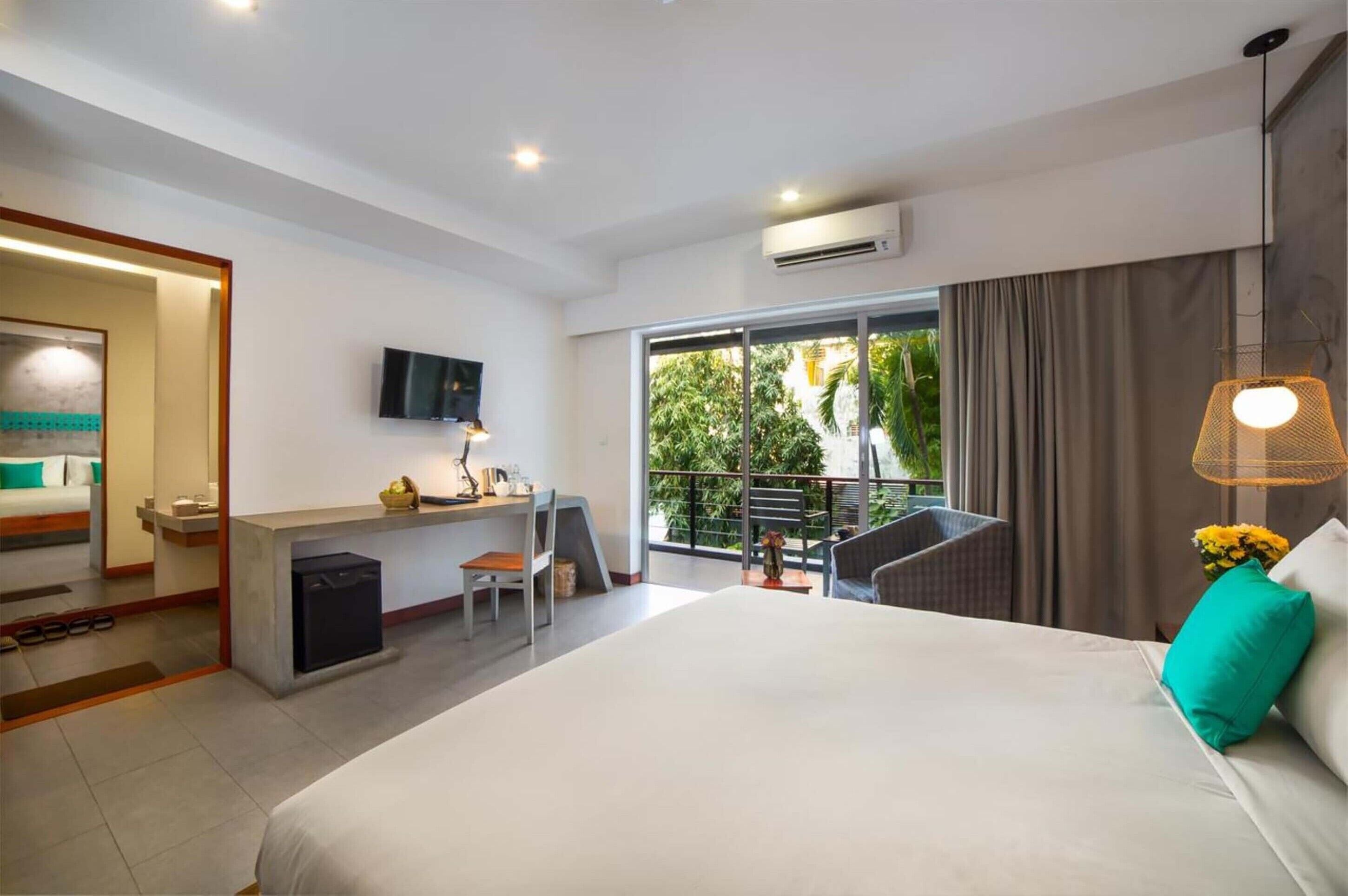 A modern hotel room with a large bed, a desk, a chair, a TV, and a balcony with a view of greenery.