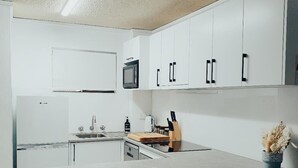 Une cuisine moderne avec des meubles blancs, un four à micro-ondes, un évier et une planche à découper en bois. On aperçoit deux tabourets en osier au premier plan.
