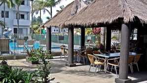 Un espace extérieur abrité, aménagé avec des tables et des chaises, une piscine en arrière-plan et une structure surmontée d’un toit de chaume.