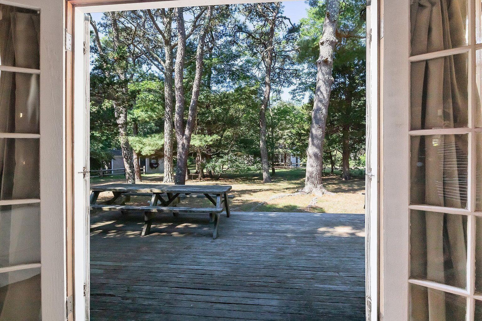 Une porte ouverte donne sur une terrasse en bois équipée de tables de pique-nique, le tout étant entouré d’arbres.