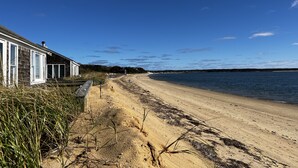 Een zandstrand met een houten huis aan de linkerkant, een heldere blauwe lucht en een rustige zee.
