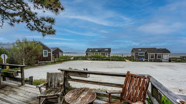 Een houten terras met een tafel en stoelen, met uitzicht op een strand met huizen op de achtergrond.