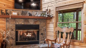 L’intérieur d’une cabane avec une cheminée en pierre, une chaise en bois portant un panneau d’accueil et une fenêtre donnant sur un paysage verdoyant.