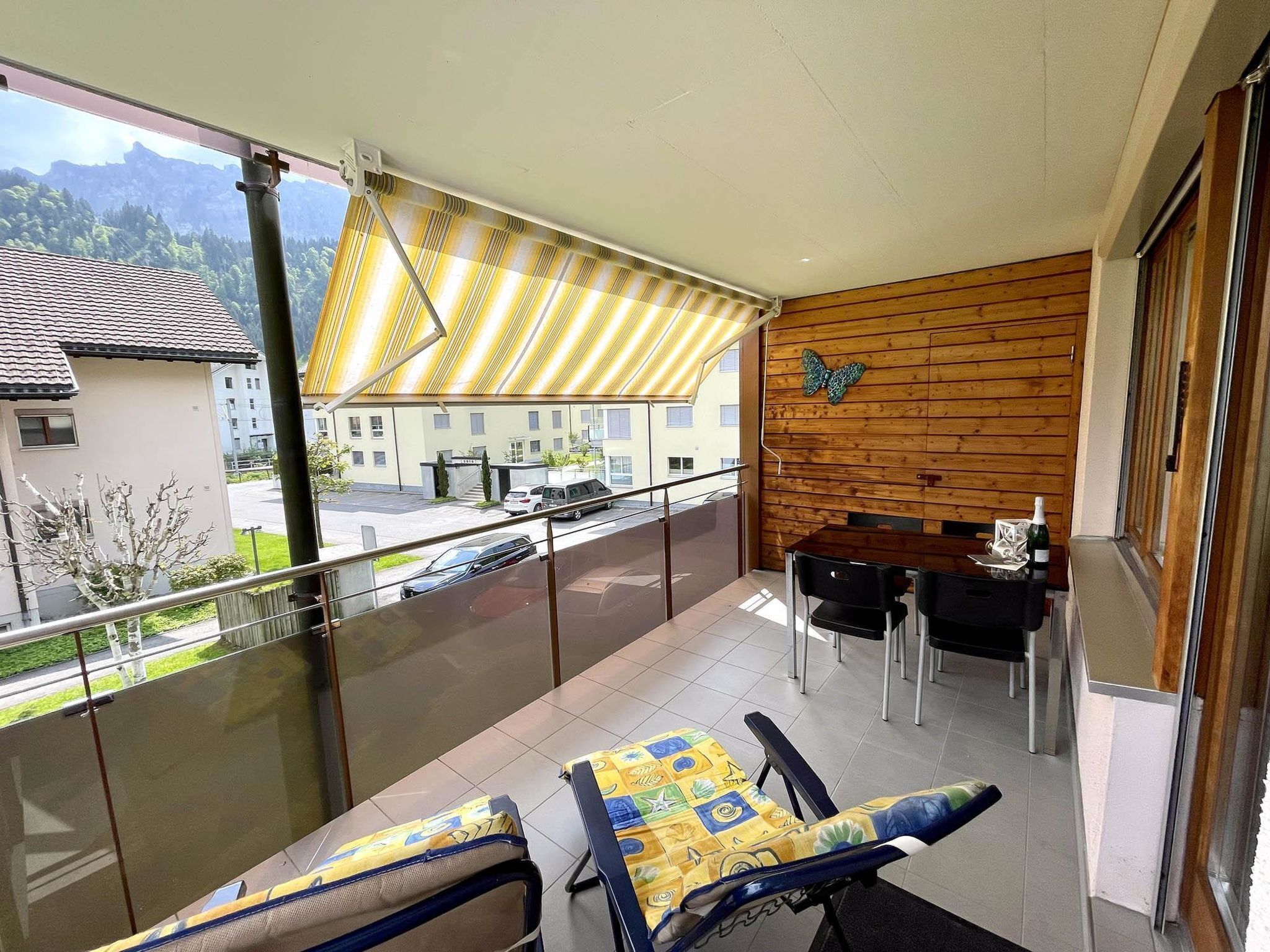 A balcony with a table and chairs, a wooden wall with a butterfly decoration, and a view of a street and buildings.