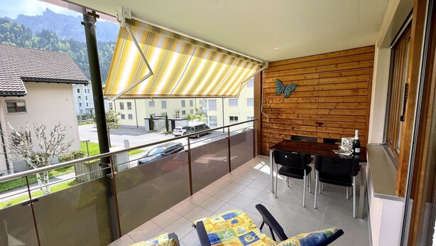Een balkon met een tafel en stoelen, een houten wand met een vlinderdecoratie, en uitzicht op een straat en gebouwen.