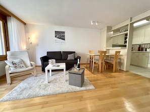Een moderne woonkamer met een bank, salontafel en stoelen, een keuken met kasten en een eettafel, en een poster aan de muur.