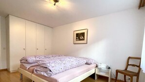 Een slaapkamer met een bed, nachtkastje, stoel, kledingkast en een ingelijst kunstwerk aan de muur.